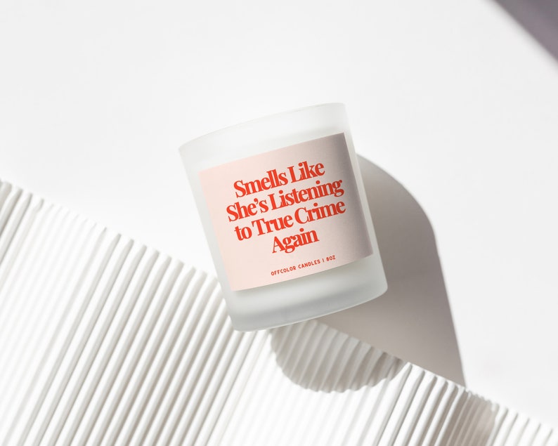 Pode incluir: Uma vela de vidro fosco com um r&oacute;tulo rosa que diz "Smells Like She's Listening to True Crime Again" em texto vermelho. A vela est&aacute; rotulada como "OFFCOLOR CANDLES | 8 OZ". A vela est&aacute; em uma superf&iacute;cie branca.