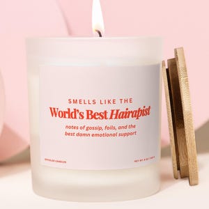 May include: A frosted glass candle with a lit wick and a bamboo lid. The label reads: SMELLS LIKE THE World's Best Hairapist, notes of gossip, foils, and the best damn emotional support. OFFCOLOR CANDLES, NET WT. 8 OZ / 244 G.