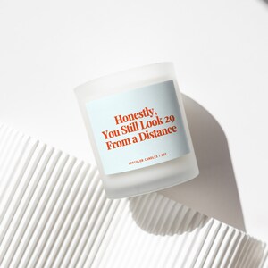 Peut inclure: Une bougie blanche avec une &eacute;tiquette bleue qui dit "Honn&ecirc;tement, tu as toujours l'air d'avoir 29 ans de loin" et "Off Color Candles | 8oz".