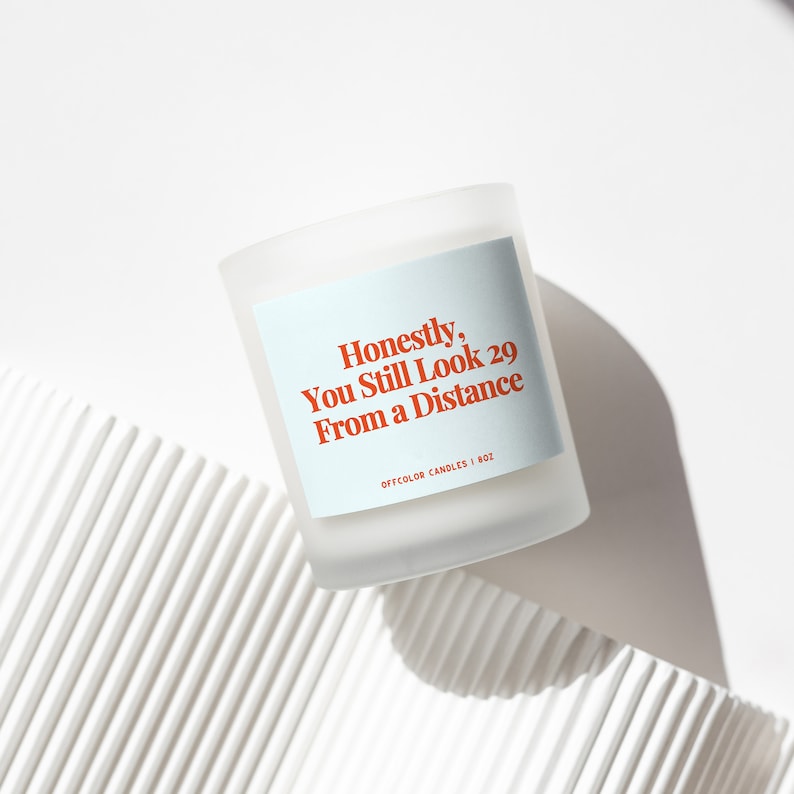 Peut inclure: Une bougie blanche avec une &eacute;tiquette bleue qui dit "Honn&ecirc;tement, tu as toujours l'air d'avoir 29 ans de loin" et "Off Color Candles | 8oz".