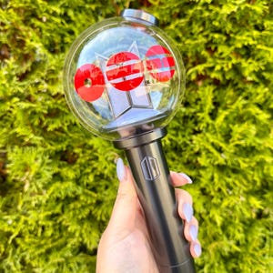 May include: A black and silver light stick with a clear, spherical top containing red and white circular designs. The handle has a silver logo. The background is a dense green bush.