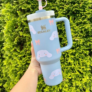 May include: A light blue stainless steel travel mug with a handle. The mug has a white cloud-shaped lid with a pink face and a pink bow. The mug is decorated with a pattern of pink bows and white cartoon dogs with pink ears and bows. The mug has the word "STANLEY" printed on the side in gold lettering.
