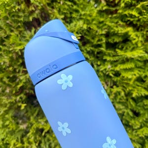 Puede incluir: Una botella de agua azul claro con tapa abatible y correa. La botella est&aacute; decorada con dise&ntilde;os de flores azul claro y la palabra "owala" est&aacute; impresa en el lateral. El fondo es un arbusto verde y frondoso.