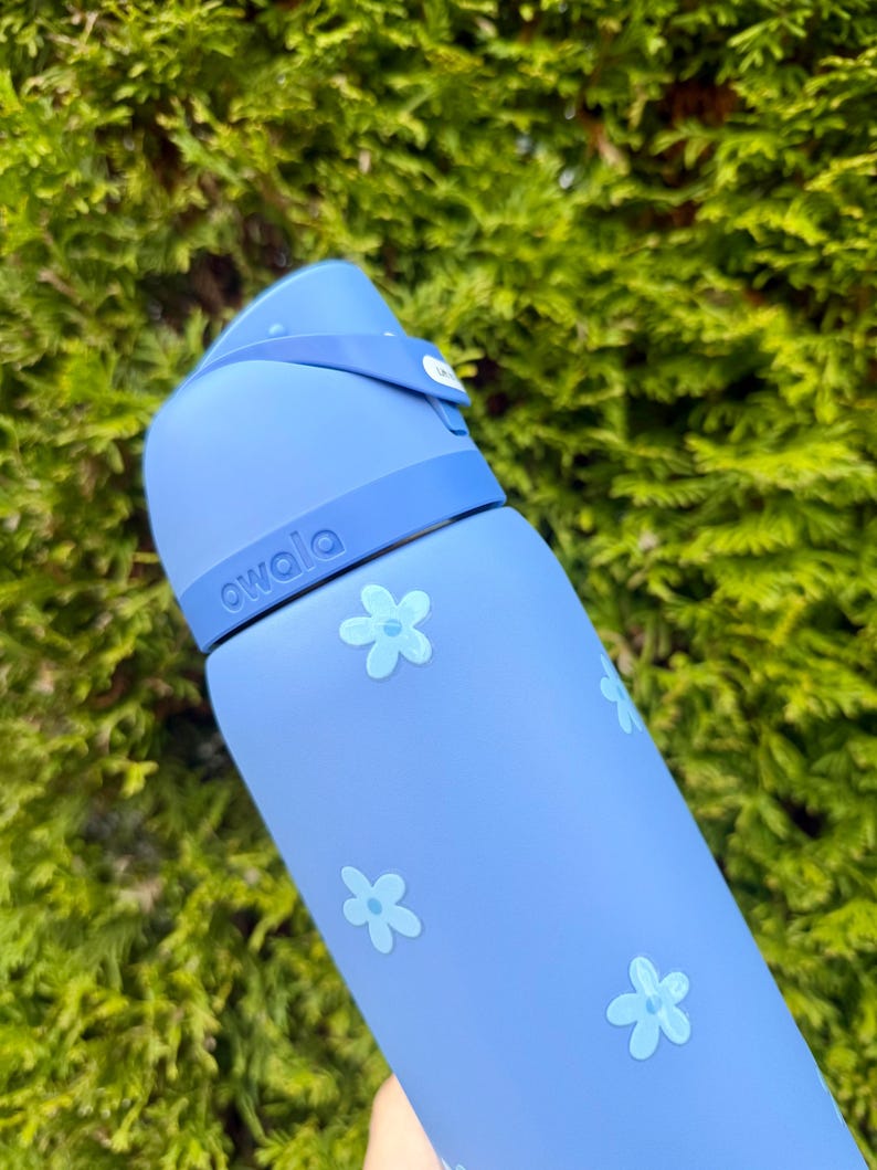 Puede incluir: Una botella de agua azul claro con tapa abatible y correa. La botella est&aacute; decorada con dise&ntilde;os de flores azul claro y la palabra "owala" est&aacute; impresa en el lateral. El fondo es un arbusto verde y frondoso.