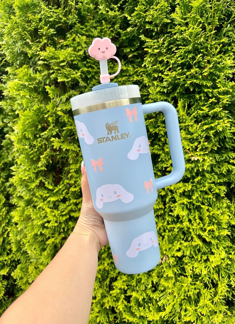 May include: A light blue stainless steel travel mug with a handle. The mug has a white cloud-shaped lid with a pink face and a pink bow. The mug is decorated with a pattern of pink bows and white cartoon dogs with pink ears and bows. The mug has the word "STANLEY" printed on the side in gold lettering.