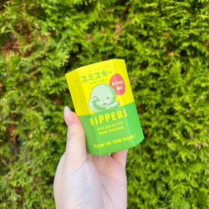 Pode incluir: Uma caixa amarela com um personagem verde na frente. A caixa diz "Smiski" e "Attach Me!" na frente. A caixa tamb&eacute;m diz "Hippers Decorative Mini Figures Glow in the Dark!"