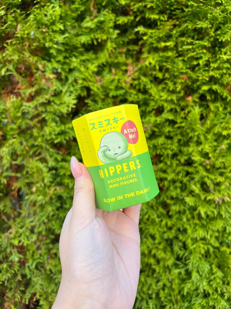 Pode incluir: Uma caixa amarela com um personagem verde na frente. A caixa diz "Smiski" e "Attach Me!" na frente. A caixa tamb&eacute;m diz "Hippers Decorative Mini Figures Glow in the Dark!"