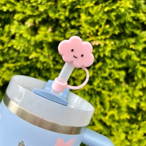 May include: A light blue stainless steel travel mug with a pink bow and a white logo that says "STANLEY". The mug has a clear lid with a pink cloud-shaped straw topper with a smiling face.