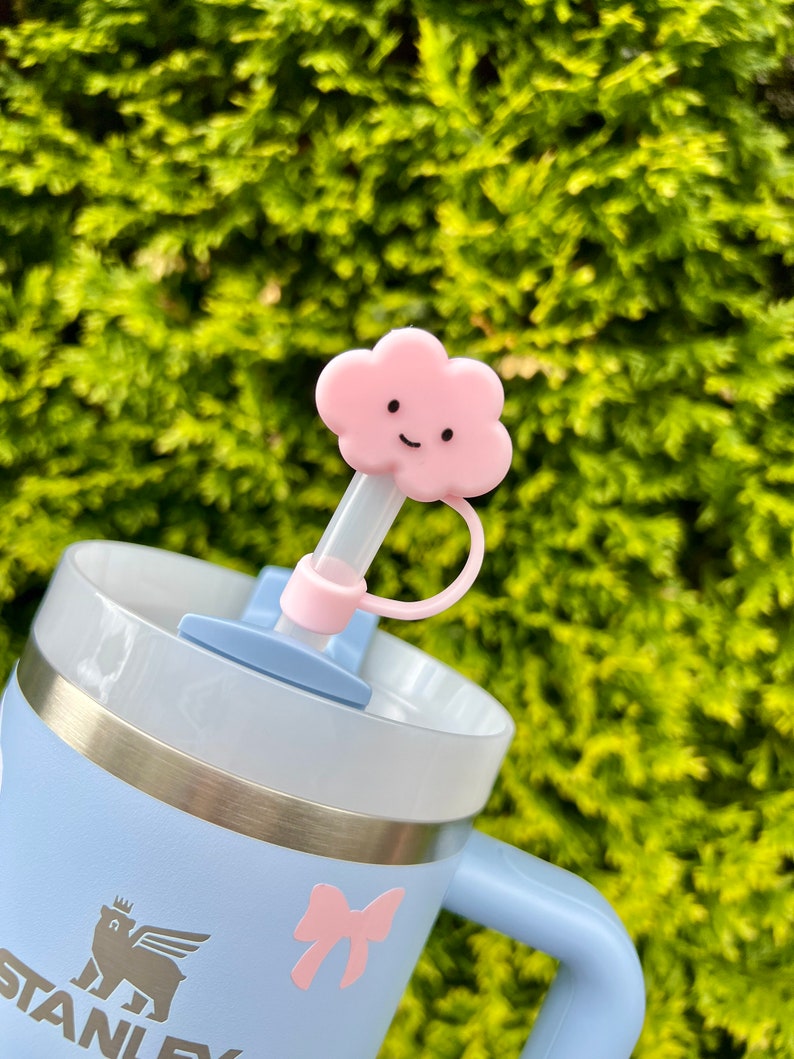May include: A light blue stainless steel travel mug with a pink bow and a white logo that says "STANLEY". The mug has a clear lid with a pink cloud-shaped straw topper with a smiling face.