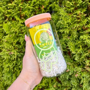 Pode incluir: Um frasco de vidro transparente com tampa rosa cheio de papel branco picado. O frasco tem um r&oacute;tulo verde e amarelo com um personagem de desenho animado e o texto "HIPPER DECORATIONS".