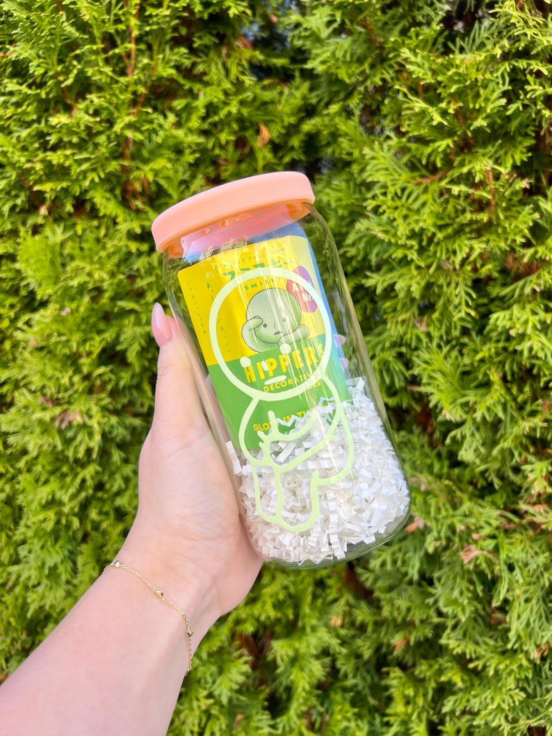 Pode incluir: Um frasco de vidro transparente com tampa rosa cheio de papel branco picado. O frasco tem um r&oacute;tulo verde e amarelo com um personagem de desenho animado e o texto "HIPPER DECORATIONS".