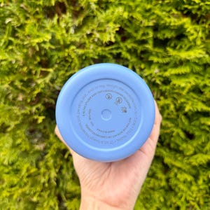 Puede incluir: Una botella de agua azul claro con base redondeada. La parte inferior de la botella tiene texto impreso, incluyendo "MADE IN CHINA" y "NOT FOR USE WITH HOT OR CARBONATED LIQUIDS". La botella se sostiene contra un fondo de follaje verde.