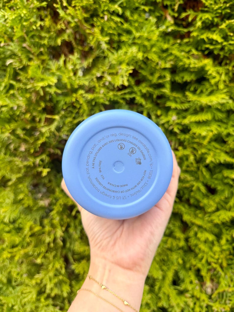 Puede incluir: Una botella de agua azul claro con base redondeada. La parte inferior de la botella tiene texto impreso, incluyendo "MADE IN CHINA" y "NOT FOR USE WITH HOT OR CARBONATED LIQUIDS". La botella se sostiene contra un fondo de follaje verde.