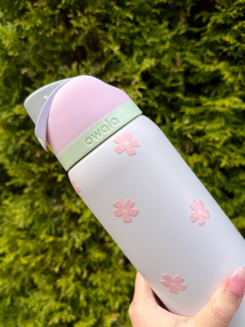Pode incluir: Uma garrafa de &aacute;gua Owala cinza claro com detalhes de flores rosa. A garrafa tem uma tampa rosa e uma faixa verde com a marca "owala". Projetada para hidrata&ccedil;&atilde;o, inclui uma al&ccedil;a para facilitar o transporte.