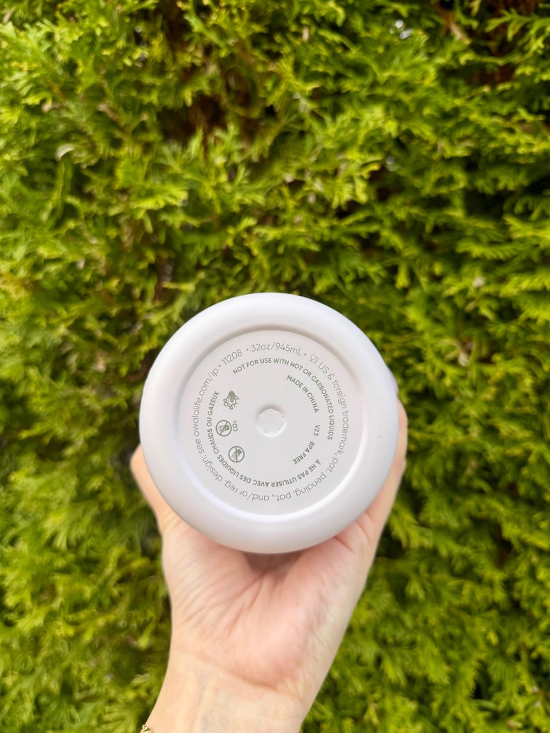 May include: A white insulated water bottle with a 32oz/945mL capacity. The bottom of the bottle has printed text including "NOT FOR USE WITH HOT OR CARBONATED LIQUIDS" and "MADE IN CHINA". The bottle is held in a hand against a green background.