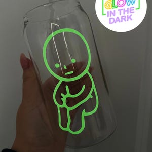 Pode incluir: Uma garrafa de &aacute;gua de vidro transparente com um desenho verde que brilha no escuro de um personagem de desenho animado. O desenho apresenta uma figura simples de cabe&ccedil;a redonda com bra&ccedil;os e pernas, e o texto "GLOW IN THE DARK" &eacute; vis&iacute;vel no fundo.