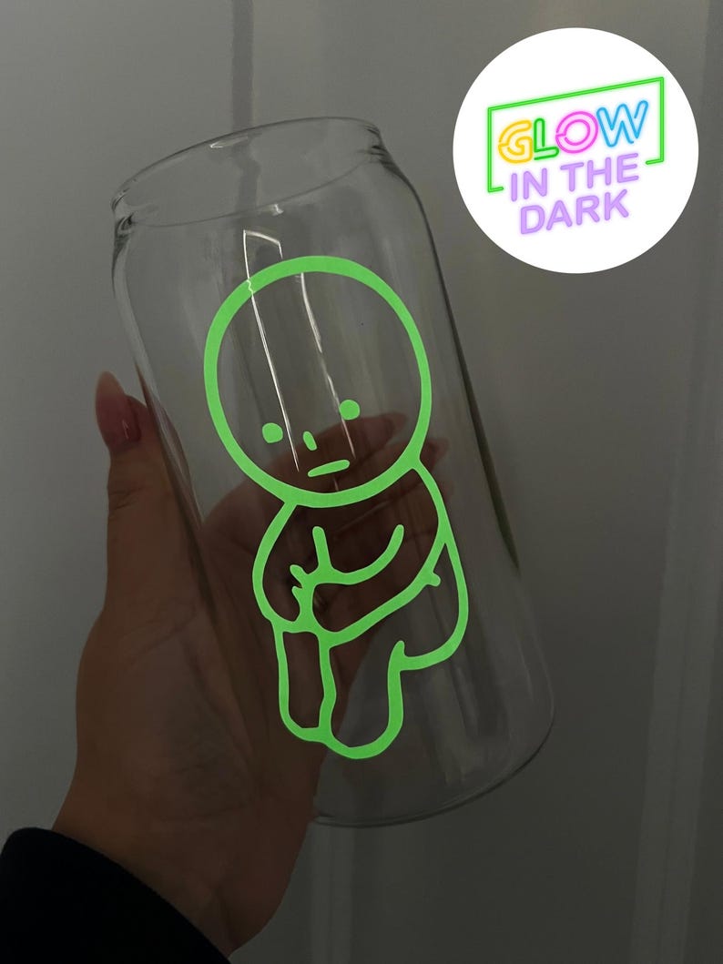 Pode incluir: Uma garrafa de &aacute;gua de vidro transparente com um desenho verde que brilha no escuro de um personagem de desenho animado. O desenho apresenta uma figura simples de cabe&ccedil;a redonda com bra&ccedil;os e pernas, e o texto "GLOW IN THE DARK" &eacute; vis&iacute;vel no fundo.