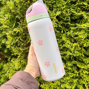 Pode incluir: Uma garrafa de &aacute;gua Owala branca com detalhes de flores cor-de-rosa. A garrafa tem uma tampa rosa e verde e uma al&ccedil;a. A palavra "owala" &eacute; impressa em dourado na garrafa. A garrafa &eacute; segurada em frente a um arbusto verde.