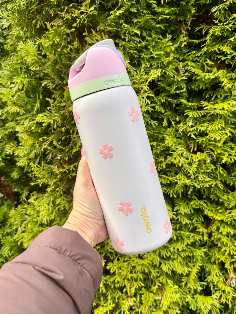 Pode incluir: Uma garrafa de &aacute;gua Owala branca com detalhes de flores cor-de-rosa. A garrafa tem uma tampa rosa e verde e uma al&ccedil;a. A palavra "owala" &eacute; impressa em dourado na garrafa. A garrafa &eacute; segurada em frente a um arbusto verde.