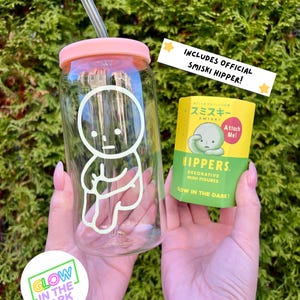 Pode incluir: Uma garrafa de &aacute;gua de vidro transparente com uma tampa rosa e um canudo. A garrafa tem um design que brilha no escuro de um pequeno personagem de desenho animado branco. A garrafa est&aacute; sendo segurada pelas m&atilde;os de uma pessoa. Tamb&eacute;m &eacute; vis&iacute;vel uma pequena embalagem amarela com um personagem que brilha no escuro. A embalagem diz "Inclui Smiski Hipper oficial!" e "Hippers. Figuras mini decorativas. Brilham no escuro!"