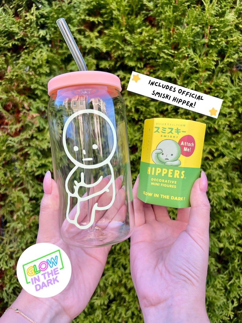 Pode incluir: Uma garrafa de &aacute;gua de vidro transparente com uma tampa rosa e um canudo. A garrafa tem um design que brilha no escuro de um pequeno personagem de desenho animado branco. A garrafa est&aacute; sendo segurada pelas m&atilde;os de uma pessoa. Tamb&eacute;m &eacute; vis&iacute;vel uma pequena embalagem amarela com um personagem que brilha no escuro. A embalagem diz "Inclui Smiski Hipper oficial!" e "Hippers. Figuras mini decorativas. Brilham no escuro!"