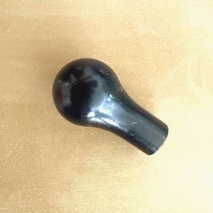 Puede incluir: Un pomo negro sólido, liso y con forma de lágrima con un acabado brillante. El objeto presenta una cabeza redondeada y bulbosa que se estrecha hacia una base cilíndrica, mostrando una superficie oscura uniforme con reflejos de luz sutiles.