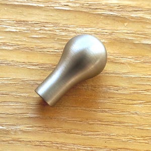 Peut inclure: Un bouton de meuble unique avec une finition argentée brossée. Le bouton présente une tête arrondie et bulbeuse qui se rétrécit vers une base cylindrique. La surface a une texture métallique subtile, reflétant la lumière sur sa forme courbe.