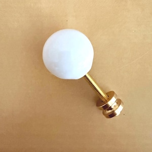 May include: A spherical white knob featuring a smooth, opaque finish attached to a gold-toned metal bolt. The hardware includes a threaded rod and a circular base plate, providing a functional component for cabinetry or furniture drawer fronts.