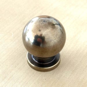Peut inclure: Un bouton de meuble sphérique de couleur bronze avec une base circulaire. Le bouton a une surface polie et réfléchissante, et la base est d'une teinte plus foncée de bronze. La quincaillerie est conçue pour être utilisée sur des tiroirs ou des armoires.