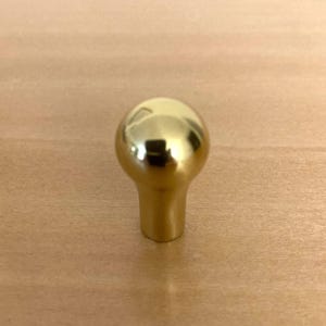 May include: A shiny, gold-colored cabinet knob with a rounded top and a cylindrical base. The knob is made of metal and reflects the surrounding light. The hardware is designed for use on drawers or cabinet doors.