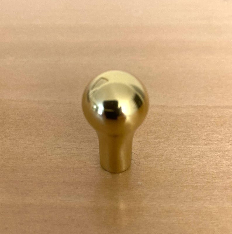 May include: A shiny, gold-colored cabinet knob with a rounded top and a cylindrical base. The knob is made of metal and reflects the surrounding light. The hardware is designed for use on drawers or cabinet doors.