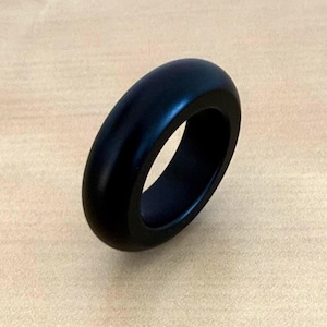 Könnte beinhalten: Ein massiver schwarzer Ring mit einem glatten, abgerundeten Design. Der Ring besteht aus einem dunklen Material und hat eine polierte Oberfläche. Der Ring ist ein schlichtes, aber elegantes Schmuckstück.