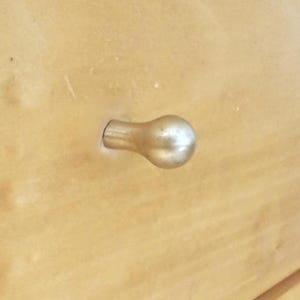 May include: Close-up of a light-colored wooden surface with a gold-colored, bulb-shaped drawer pull. The drawer pull is attached to the wood with a short, cylindrical stem.