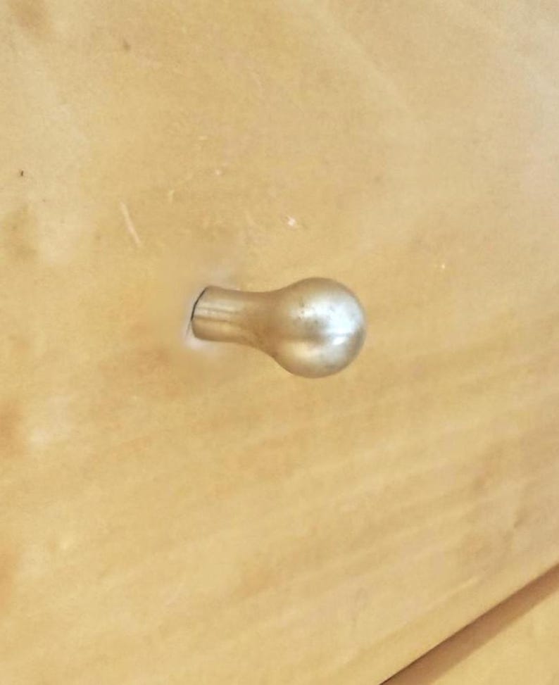 May include: Close-up of a light-colored wooden surface with a gold-colored, bulb-shaped drawer pull. The drawer pull is attached to the wood with a short, cylindrical stem.