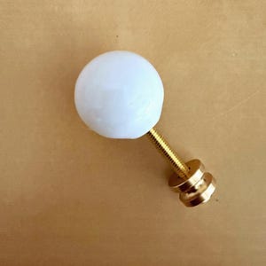 May include: A white, spherical knob with a brass-colored threaded stem and base. The knob is likely for a cabinet, drawer, or other furniture piece. The hardware is set against a neutral background.