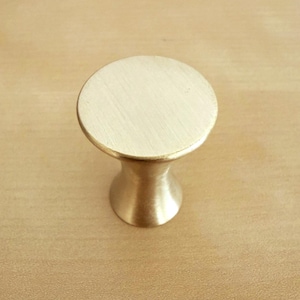 May include: A circular cabinet knob with a brushed gold finish. The knob features a flat, round top surface that transitions into a tapered, hourglass-shaped base. The metal surface has a subtle, fine-grained texture visible across the top.