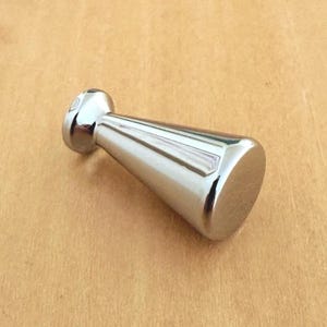 以下が含まれることがあります： 先細りの円錐形をした、磨き上げられたシルバー調の金属製家具用ノブ。金具は光を反射する滑らかな表面を持ち、円形のベースと、より幅の広い平らな上端部を備えており、引き出しや家具に取り付けるためのデザインとなっています。
