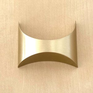 Puede incluir: Un tirador de mueble de metal dorado cepillado con un diseño curvo y cóncavo. El herraje presenta un acabado metálico liso con una forma distintiva de reloj de arena, caracterizada por lados curvados hacia adentro que crean un perfil arqueado.