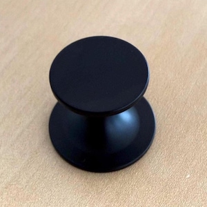 Op de afbeelding: Zwarte kastknop met een ronde boven- en onderkant, verbonden door een gebogen steel. De knop heeft een matte afwerking en staat op een lichtbruin oppervlak. Geschikt voor laden en kasten.