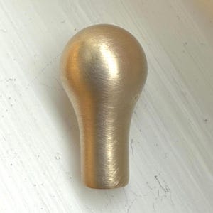 Könnte beinhalten: Ein Möbelknopf aus massivem Messing mit abgerundeter Oberseite und zylindrischem Sockel. Der Knopf hat eine gebürstete Oberfläche, die ihm ein leicht strukturiertes Aussehen verleiht. Die Hardware ist für Schubladen oder Schranktüren konzipiert.