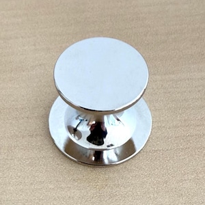 May include: A shiny, silver-colored cabinet knob with a circular top and base. The knob has a rounded, bulbous midsection and reflects the surrounding environment. The knob is likely made of metal and is designed for use on furniture.