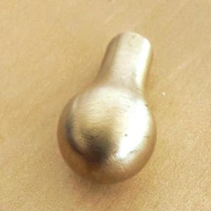 May include: A brass-colored, pear-shaped cabinet knob with a rounded base and a rectangular top. The knob has a smooth, polished surface and is likely used for drawers or doors. The hardware is shown against a light brown background.