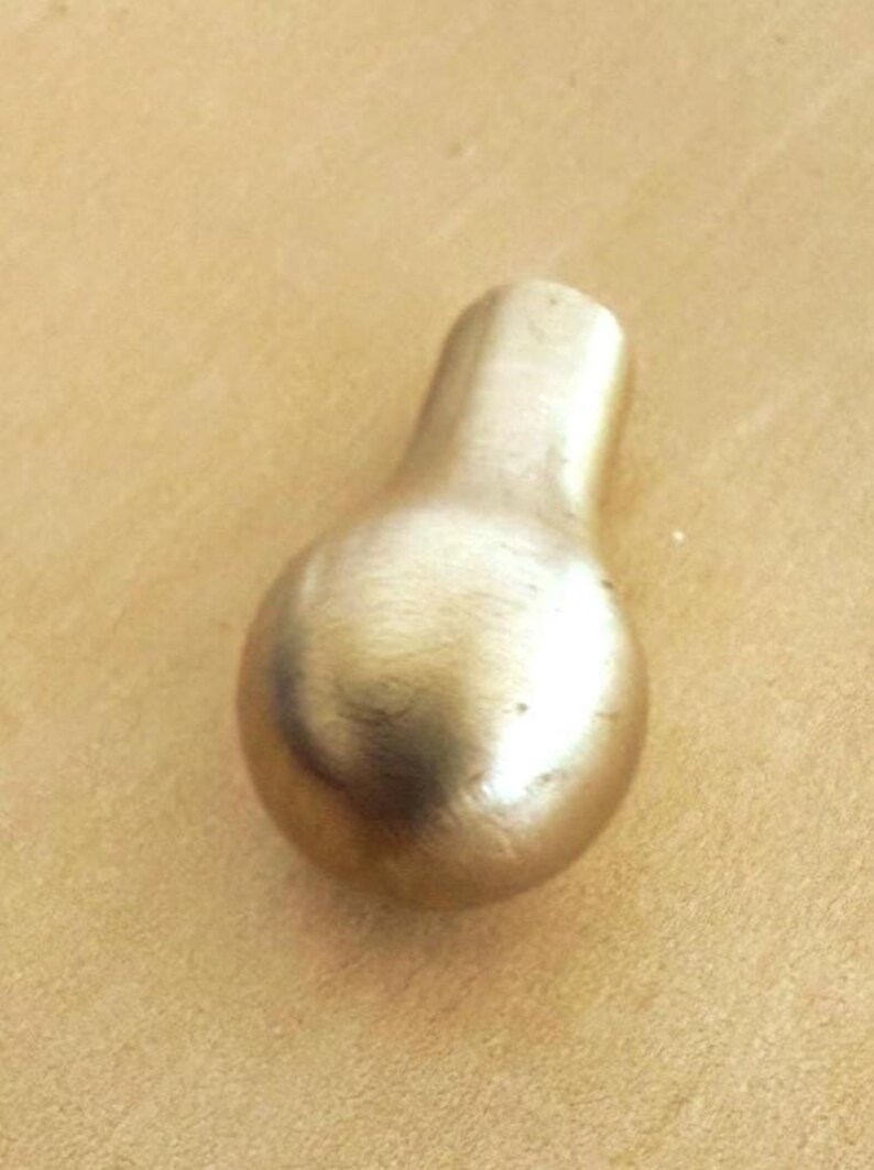 May include: A brass-colored, pear-shaped cabinet knob with a rounded base and a rectangular top. The knob has a smooth, polished surface and is likely used for drawers or doors. The hardware is shown against a light brown background.