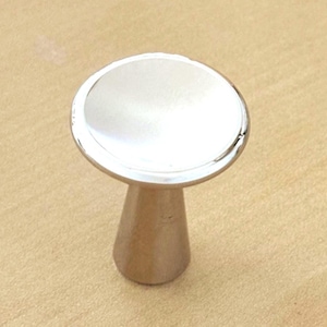 Könnte beinhalten: Ein silberfarbener Möbelknopf aus Metall mit einer glatten, kreisförmigen, leicht konkaven Oberseite und einem konisch zulaufenden, zylindrischen Sockel. Das Beschlagteil hat eine polierte, reflektierende Oberfläche, die das Licht einfängt.