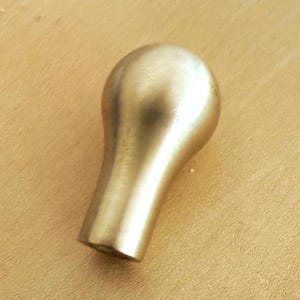 May include: A solid brass cabinet knob with a smooth, rounded, teardrop-shaped head and a cylindrical base. The metal surface has a soft, brushed gold-toned finish that reflects light evenly across its curved, polished exterior form.