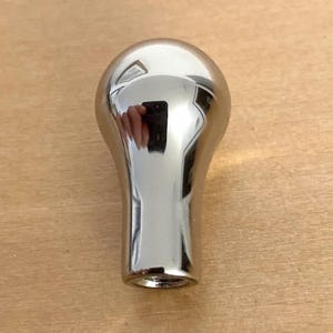 May include: A chrome-finished, teardrop-shaped knob. The smooth, reflective surface of the knob mirrors the surrounding environment. The knob has a rounded top and a cylindrical base. The object is likely a furniture or appliance component.
