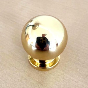 May include: A shiny, gold-colored cabinet knob with a spherical top and a circular base. The knob reflects the surrounding environment, creating a mirrored effect. The hardware is designed for furniture and home decor.