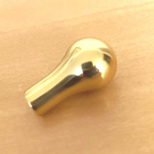 May include: A small, teardrop-shaped cabinet knob with a polished, reflective gold-tone finish. The object features a smooth, rounded head that tapers down into a cylindrical base, creating a simple, solid, and metallic hardware component.