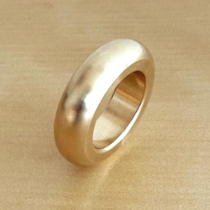 Puede incluir: Un anillo de tono dorado pulido con una banda gruesa y redondeada, que presenta una superficie lisa y reflectante. El anillo tiene un diseño circular simple y continuo con un perfil ancho, mostrando un brillo metálico cálido y uniforme.