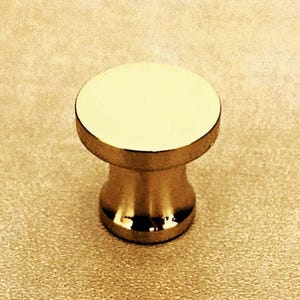 Puede incluir: Un pomo para mueble de color dorado con una parte superior circular plana y lisa y una base cilíndrica cónica. El herraje presenta un acabado metálico pulido y reflectante que capta la luz, creando un aspecto limpio y uniforme.